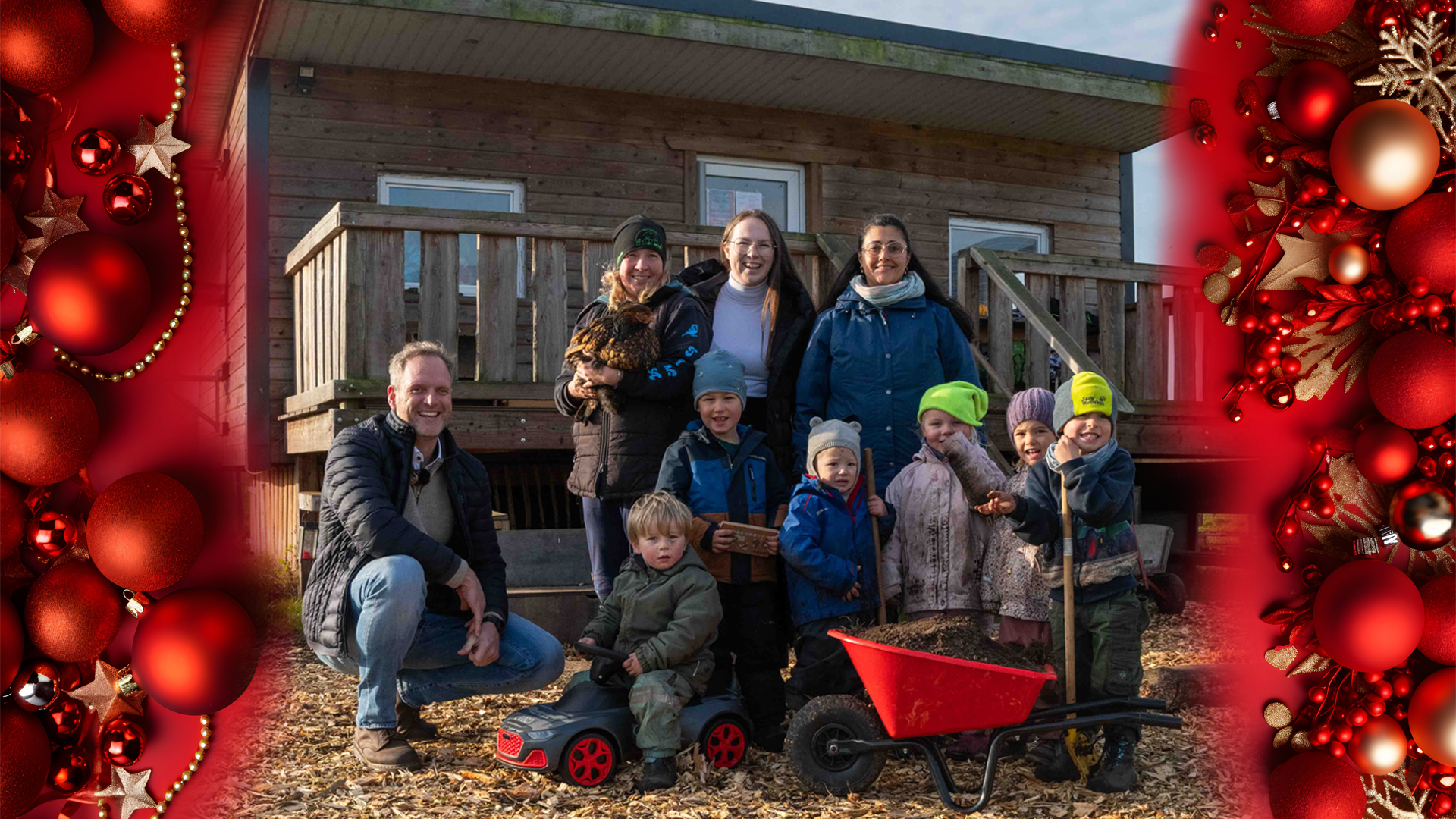 Mehrere Erwachsene und Kinder posieren vor einem kleinen Holzhaus auf einem ländlichen Gelände. Einige Kinder halten Spielzeug oder Werkzeuge wie Schubkarre und Laufrad, eine Person trägt ein Huhn. Alle blicken lächelnd in die Kamera.